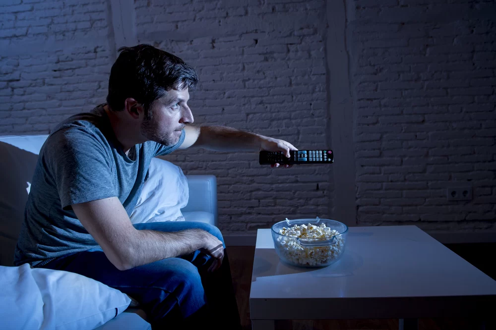 Man watching TV late at night with bowl of popcorn on table and remote in hand