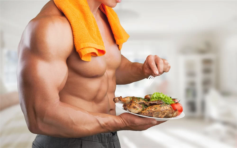 Muscled shirtless man with towel draped over neck holding a plate of chicken and salad