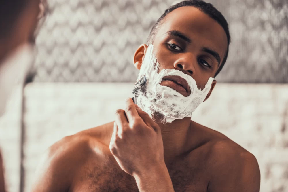 Young African American man shaving his face with razor and shaving cream