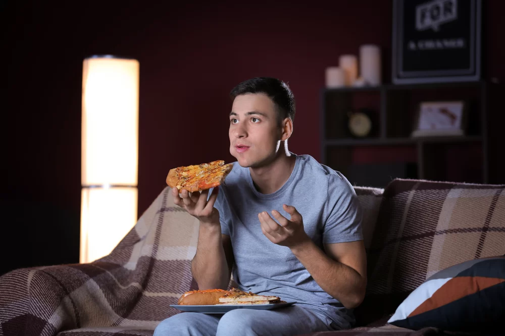 Man eating large slice of pizza while watching TV on the couch