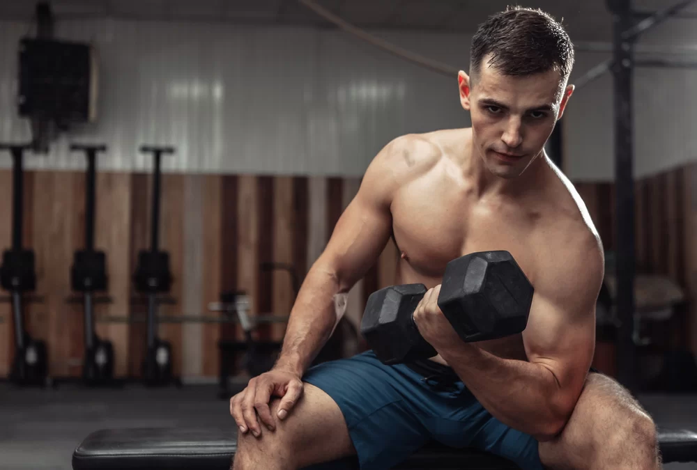 Fit, shirtless man sitting on bench doing bicep curls in gym