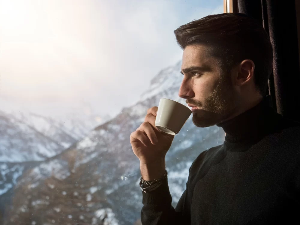 Man sipping from mug while staring out window at snow-covered mountains