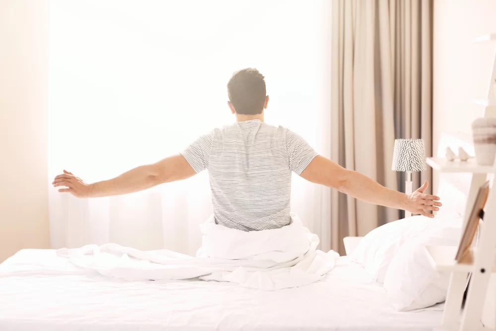 Man stretching both arms out while sitting up in bed after just waking up